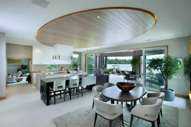 Bright, open-plan contemporary kitchen, dining, and living area with a large island, integrated appliances, white cabinetry, and a round dining table in the foreground. A distinctive curved timber-lined ceiling feature with a warm timber edge spans the room. Full-height sliding glazing opens to an outdoor entertaining area and water view, while indoor plants and neutral furnishings create an airy, refined atmosphere.
