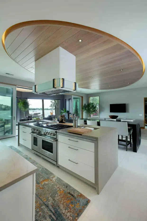 Contemporary open-plan kitchen with a large central island, stainless-steel range cooker and hood, pale fixed cabinetry, and a light stone benchtop. A dramatic curved timber-panel ceiling feature spans the kitchen and dining area, with large glazed openings connecting to an outdoor view. The adjoining dining space includes a dark dining table, white upholstered chairs, and a wall-mounted television. The room is bright, refined, and open, with indoor plants and a patterned runner adding warmth.
