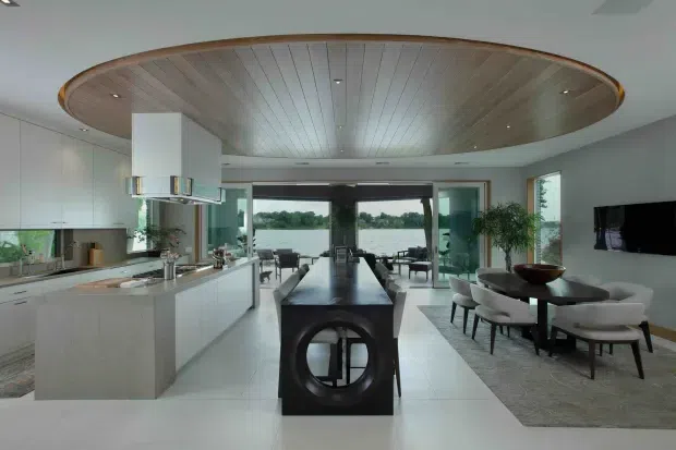 Open-plan luxury kitchen, dining and living area with a large sculptural island beneath a distinctive oval timber-lined ceiling feature. White fixed cabinetry and pale flooring create a bright, refined interior, while full-width glazing opens toward a waterfront view. A dining setting and lounge area are visible beyond the kitchen.