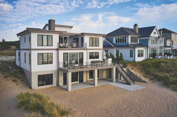 Exterior view of a contemporary multi-level coastal residence set directly among sandy dunes, with broad glazing, balconies, terraces, and a lower level opening toward the beach. Traditional neighbouring homes are visible behind it under a partly cloudy sky.