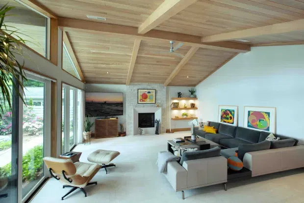 A bright, spacious living room with floor-to-ceiling glazing along one side, a warm timber-lined pitched ceiling with exposed beams, pale flooring, and a contemporary sectional seating area arranged around a low coffee table. A fireplace, floating timber shelves with decor, colourful artwork, and a lounge chair add character. Garden planting is visible through the glazing.