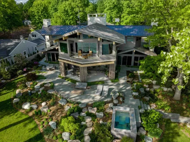 Aerial exterior view of a large, multi-level residential house set within a mature, heavily landscaped garden. The home features multiple gabled roof forms with dark metal roofing, broad roof overhangs, stone-clad structural columns, extensive glazing, upper-level balconies, and covered outdoor living areas. Paved paths, boulders, planting beds, lawn, and a small rectangular water feature or plunge pool surround the building.