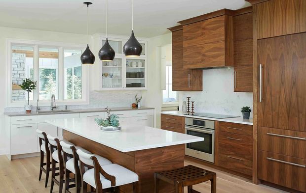 Bright, spacious contemporary kitchen with a large white island and integrated seating for four. The room features rich walnut-toned timber cabinetry, white stone-look benchtops, a pale marble-look splashback, pendant lighting, open shelving, and generous windows overlooking greenery. Dark wood bar stools and a small timber stool add warmth to the refined neutral scheme.