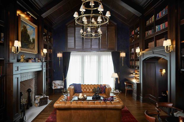 A richly furnished, dark-toned library or study with floor-to-ceiling timber bookcases, a coffered or exposed dark timber ceiling, a central leather sofa and ottoman, a traditional fireplace with pale mantel, and multiple brass chandelier fixtures. Plantation shutters and sheer curtains cover the tall central window, while framed artwork, books, and classic table and wall lighting create a warm, formal atmosphere.