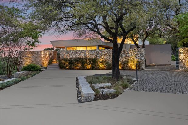 A single-storey contemporary residence viewed from the front at dusk, with a broad smooth concrete driveway and pedestrian approach leading to a recessed entry. The property is enclosed by illuminated dry-stone masonry walls, with a flat-roofed home and warm horizontal glazing partially visible behind them. Mature trees, gravel paving, planted beds, and integrated landscape lighting create a refined, low-key exterior setting.