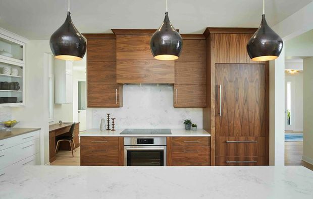 A refined contemporary kitchen with extensive dark walnut-toned fixed cabinetry, a central cooking zone, integrated refrigerator cabinetry, pale stone benchtops and splashback, and three large dark pendant lights. A white island dominates the foreground, with a small adjoining dining or workspace visible to the left.