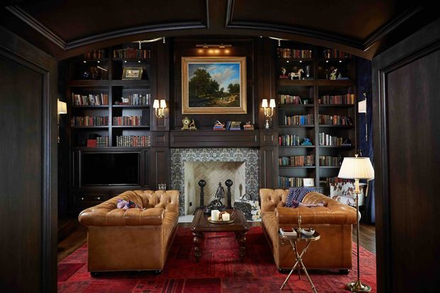 A dark, richly furnished library-style living room with floor-to-ceiling built-in bookshelves, a central traditional fireplace, framed artwork, and a television integrated into the joinery. Two cognac leather sofas face each other across a wooden coffee table, with a red patterned rug, brass lamps, and dark timber wall and ceiling panelling creating a warm, intimate atmosphere.