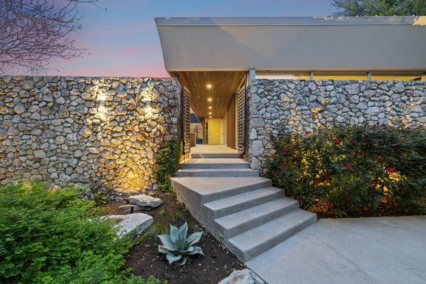Exterior dusk view of a low-slung contemporary building entrance, framed by substantial irregular natural-stone walls. A broad concrete stairway leads to a recessed, warmly lit entry corridor beneath a projecting flat roof. Desert-style planting and shrubs border the concrete approach, while integrated wall lighting highlights the stone texture.