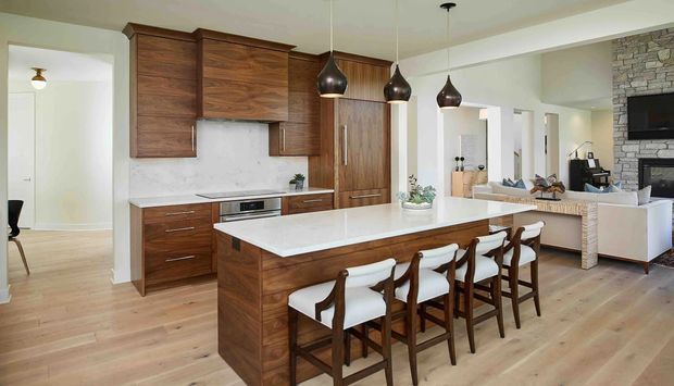 Open-plan kitchen with a large white island and seating for five, featuring rich dark timber cabinetry, pale timber flooring, white benchtops, and dark sculptural pendant lights. A living area with a stone-clad fireplace and television is visible beyond the kitchen.