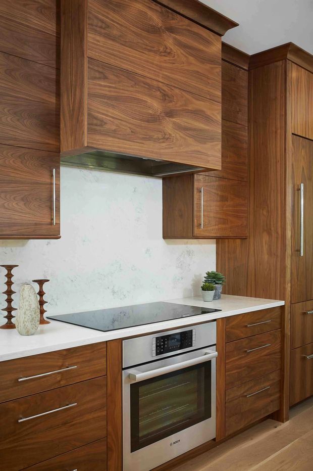 A compact, richly finished kitchen featuring extensive dark walnut-grain cabinetry, a white stone benchtop and matching light backsplash, black induction cooktop, stainless-steel built-in oven, and tall pantry-style joinery. The cabinetry includes a prominent overhead rangehood enclosure and long brushed metal handles. Timber flooring and a few small decorative objects complete the warm, refined space.