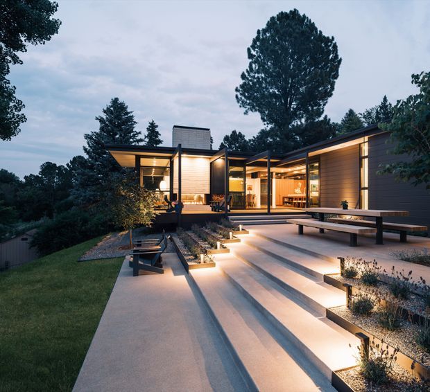 Twilight exterior view of a low-slung contemporary home integrated into a wooded, sloping site. The house features extensive floor-to-ceiling glazing, dark horizontal cladding, flat roof planes, warm timber-lined interiors, and broad concrete steps leading to a landscaped terrace with built-in bench seating and subtle step lighting.