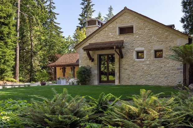 Exterior view of a rustic stone house set within a mature, heavily wooded landscape. The main gabled structure has cream-colored rough-cut stone walls, dark timber-framed glazed entry doors, small square windows, a projecting timber entry canopy, and a tiled pitched roof with a small roof dormer or cupola. A lower adjoining wing with a terracotta-toned tiled roof is visible to the left, while ferns and lawn occupy the foreground.