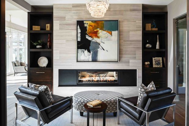 A refined living room centred on a contemporary fireplace wall clad in pale horizontal timber-look panels. Dark built-in shelving flanks the fireplace and displays decorative objects, while two black leather armchairs face a patterned upholstered ottoman and small round coffee table. A sculptural crystal chandelier, abstract artwork and large glazed openings add a polished, luxurious feel.