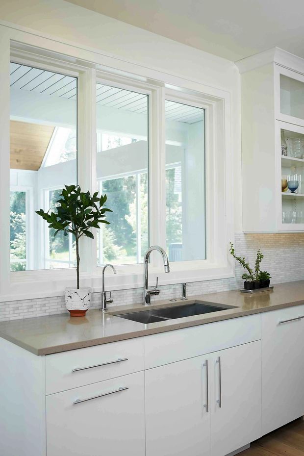 Bright, light-filled contemporary kitchen with a long white island or base-cabinet run beneath large white-framed windows. The kitchen features a pale stone benchtop, stainless steel undermount sink with twin taps, white cabinetry with simple linear handles, and a small light brick mosaic splashback. Glazed display cabinetry with glassware is visible at right, while timber flooring and greenery add warmth.