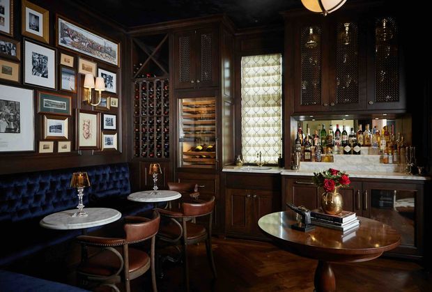 A richly detailed, dark-toned home bar with extensive dark timber cabinetry, glass-fronted upper cupboards, integrated wine storage, a mirrored or glass-backed liquor display, and a marble-look bar counter. The room includes a tufted banquette, small round marble-top café tables, leather dining chairs, framed artwork, brass wall sconces, and a central round timber table. A patterned leaded-glass window and herringbone timber flooring add traditional character. The atmosphere is intimate, luxurious, and club-like.