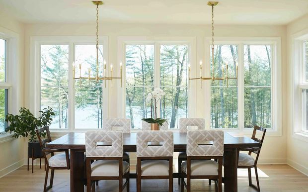 Bright dining room with a long dark timber dining table and upholstered dining chairs, surrounded by large white-trimmed windows overlooking a wooded landscape. Two linear brass-toned pendant chandeliers hang above the table, with a potted plant and orchid adding natural detail. The room has a light, refined, traditional feel.