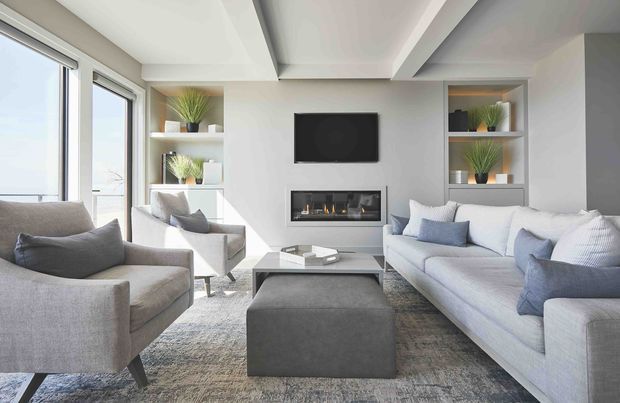 Bright, contemporary living room with a large sectional sofa, armchairs, ottoman, coffee table, wall-mounted television and linear built-in fireplace. White built-in shelving with decorative plants frames the fireplace wall, while expansive glazing provides an airy outlook. The room uses a soft palette of white, light grey and muted blue with a low-pile textured rug and streamlined furnishings.