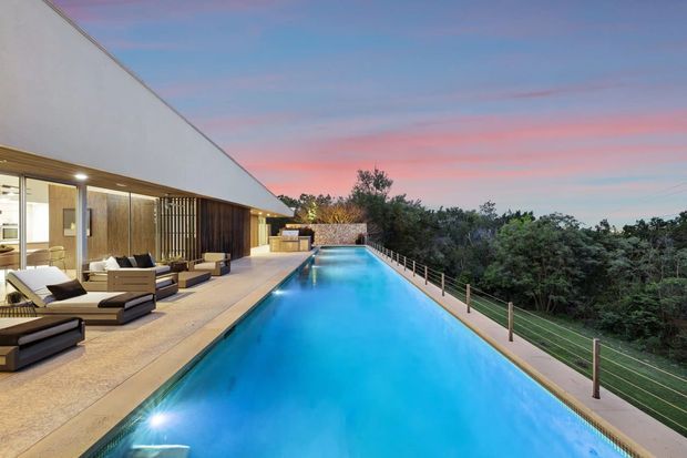 A modern single-storey residence viewed from an expansive elevated pool terrace at dusk. The long rectangular swimming pool dominates the foreground, bordered by a pale paved deck and a slim metal-and-glass balustrade overlooking dense trees. The house features a crisp white angular roof or wall plane, extensive floor-to-ceiling glazing, warm timber screening and cladding, and integrated outdoor lounge furniture. The scene has a refined contemporary resort-like character with vivid turquoise pool water and a pink-blue sunset sky.