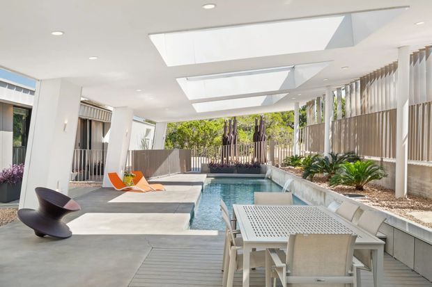 A bright, contemporary covered outdoor entertaining area beside a narrow swimming pool. The space features a concrete floor, timber decking, large skylights, white structural columns, vertical screening, and a dining setting overlooking the pool. Tropical planting and privacy fencing frame the far end, while sculptural outdoor seating and sun loungers add a relaxed resort-like character.