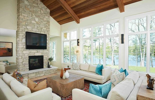 Bright, spacious living room with a large stone-clad fireplace wall, wall-mounted television, exposed timber beam ceiling, extensive white-framed glazing, and multiple cream sofas arranged around a substantial timber coffee table. The windows overlook trees and a body of water, while colourful cushions and a patterned rug add accents.