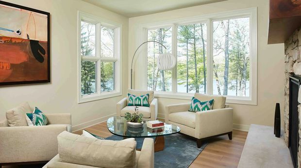 Bright, inviting living room with a corner arrangement of large white-framed windows overlooking trees, pale upholstered armchairs and sofa, a glass coffee table, patterned teal cushions, timber flooring, and a stone fireplace at right. An abstract artwork and sculptural floor lamp add restrained decorative accents.
