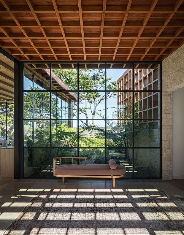 A refined interior lounge or living area with a low upholstered bench-style sofa positioned in front of expansive black-framed, grid-patterned glazing. The glazing overlooks a lush landscaped courtyard and reflects surrounding timber-clad architecture. A richly stained timber coffered ceiling, pale textured masonry wall, dark tiled floor, and strong geometric sunlight shadows create a warm, calm, indoor-outdoor atmosphere.