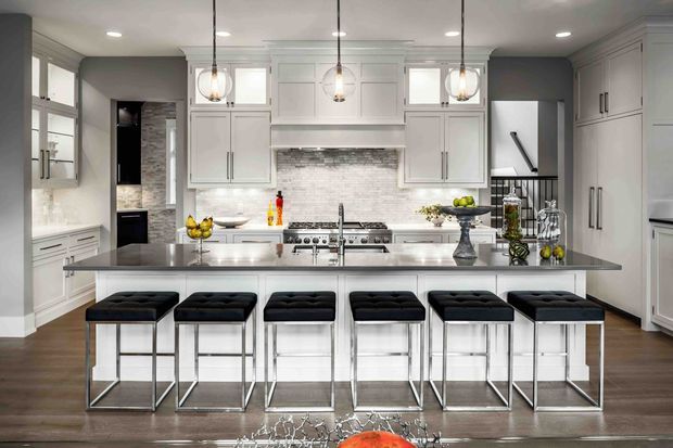 A spacious, bright kitchen dominated by a large central island with a grey stone benchtop and six black upholstered bar stools. White shaker-style cabinetry spans the walls, with integrated appliances, a stainless-steel range and hood surround, and a pale textured tile backsplash. Three pendant lights hang above the island, while dark timber flooring and stainless-steel accents provide contrast.