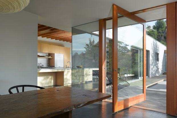 Interior view of a dining area opening onto an outdoor deck through a large timber-framed glazed door. A substantial timber dining table dominates the foreground, while a compact kitchen with light cabinetry and stainless-steel surfaces is visible behind a glass partition. Exposed timber ceiling beams and extensive glazing create a warm, indoor-outdoor connection.