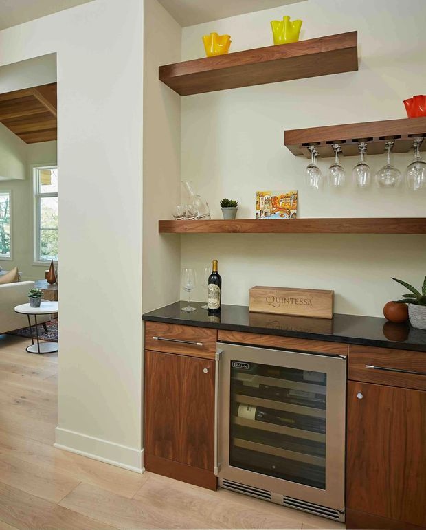 A compact built-in home bar or wine storage area with dark walnut cabinetry, a black stone benchtop, open walnut display shelves, and an under-counter glass-front wine refrigerator. Stemware hangs beneath one shelf, while bottles, glasses, and decorative objects are arranged on the counter and shelves. The space opens toward a light-filled living area with timber flooring and white walls.