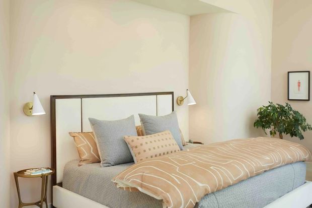 A softly lit bedroom with a neatly made bed featuring a dark timber-framed headboard, pale blue-grey bedding, and a beige patterned duvet. Wall-mounted brass-and-white sconces flank the bed, while a small round side table, leafy indoor plant, and framed artwork add warmth. The room has simple painted walls and a calm, understated contemporary interior.