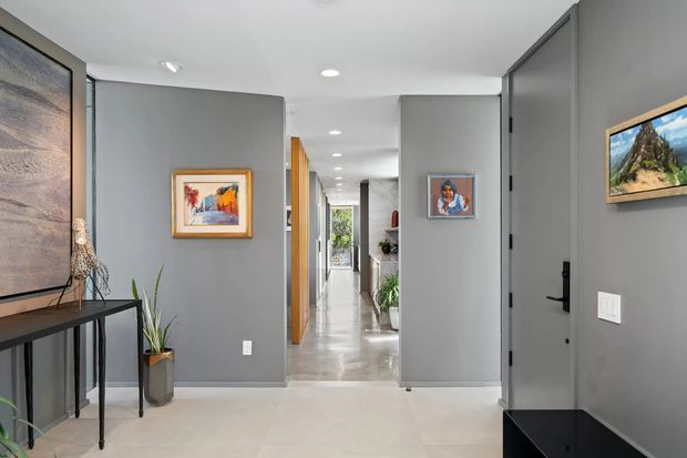 A contemporary residential entryway opening into a long, bright hallway. The space has smooth grey walls, pale polished flooring, a dark grey front door with black hardware, recessed ceiling lights, timber-trimmed doorways, framed artwork, a slim black console, and indoor plants. Glazing or an opening at the far end brings in natural light and greenery.