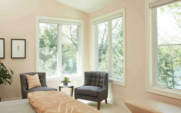 Bright, calm sitting nook with two grey tufted armchairs, a small wooden side table, and a low upholstered ottoman or bench. The room has warm beige walls, white trim, multiple large windows overlooking greenery, framed artwork, and light neutral furnishings.