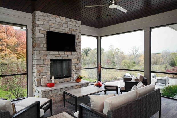 Enclosed sunroom or screened living area with expansive floor-to-ceiling glazing, a substantial natural-stone fireplace wall with built-in television, dark timber plank ceiling, ceiling fan, and woven outdoor-style seating arranged around a coffee table. The room overlooks a wooded landscape and is bright, airy, and connected to the outdoors.