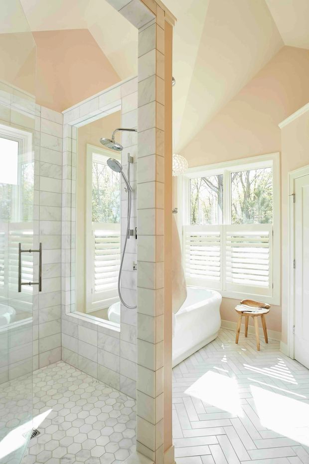 Bright, airy bathroom with a tiled walk-in shower in the foreground and a freestanding bathtub positioned beside large windows with white plantation shutters. The room features pale marble-look wall tiles, small hexagonal shower-floor tiles, light herringbone floor tiling, white fixtures, and a central tiled partition column. Natural light creates a clean, serene atmosphere.