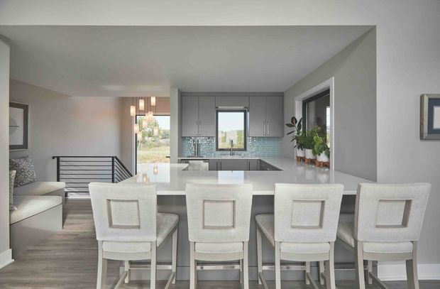 Bright open-plan kitchen and living area viewed from the dining side, featuring a large white island with four upholstered bar stools, pale grey cabinetry, a light blue glass-tile splashback, stainless-steel appliances, and grey timber-look flooring. A lounge area and stair railing are visible to the left, with large windows bringing in natural light.