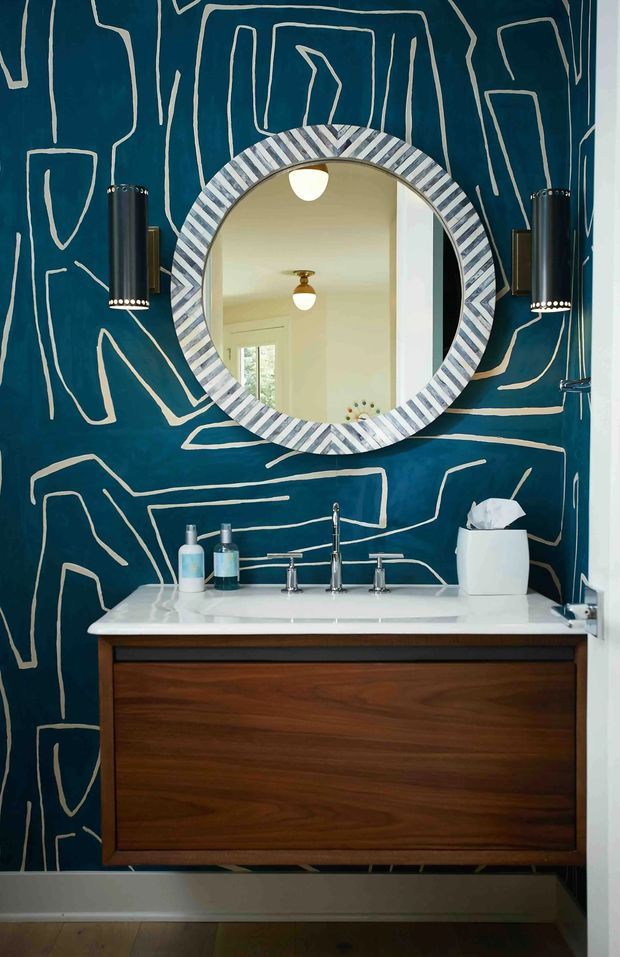 A compact, dramatically styled bathroom vanity area. A floating dark timber vanity supports a white integrated basin with chrome tapware. The wall behind is covered in deep teal wallpaper with an abstract cream line pattern, complemented by a round mirror with a bold striped blue-grey frame and two black cylindrical wall sconces. A small window is visible in the mirror reflection, and the room has timber flooring and white trim.