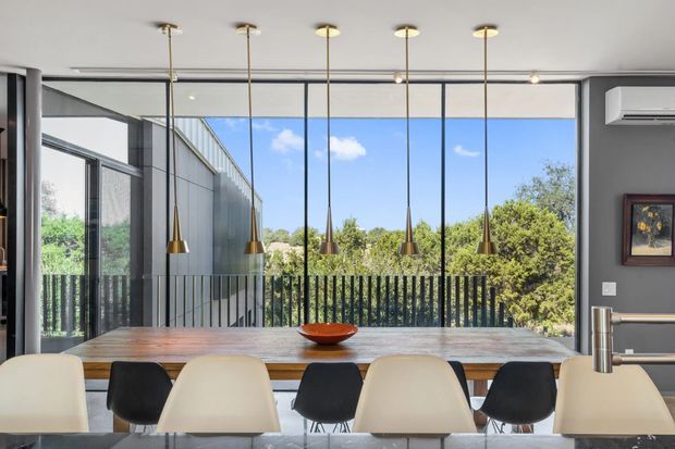 Contemporary dining area with a long timber dining table and mixed cream and black chairs, positioned in front of expansive floor-to-ceiling glazing. A row of slim brass pendant lights hangs above the table, while dark grey walls, black-framed glazing, and a stainless steel tap or fixture contribute to the refined modern interior. Lush trees and a neighbouring structure are visible beyond the glazing.