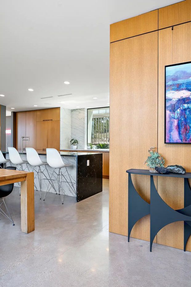 A bright, open-plan contemporary kitchen and dining area with extensive warm timber wall panelling and integrated cabinetry. A marble-look island with dark stone sides is paired with white bar stools, while polished concrete flooring extends through the space. Recessed ceiling lights, a timber dining table, framed artwork, and a sculptural dark console complete the refined interior.