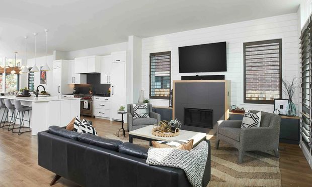 Open-plan contemporary living and kitchen area with a black leather sofa, grey armchairs, a central fireplace and wall-mounted television. The white kitchen includes an island with bar seating, pendant lights, white cabinetry and dark benchtops. Timber flooring, horizontal wall panelling, patterned window coverings and neutral furnishings create a bright, refined interior.