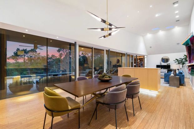 A spacious contemporary open-plan dining and living area with a large timber dining table surrounded by upholstered chairs. Floor-to-ceiling glazed doors line one side of the room, opening views to landscaped surroundings. The space has pale timber flooring, white walls and ceiling, recessed lighting, a sculptural linear pendant, and a minimalist kitchen or bar joinery zone with warm wood cabinetry. A living area and shelving are visible beyond the dining space.