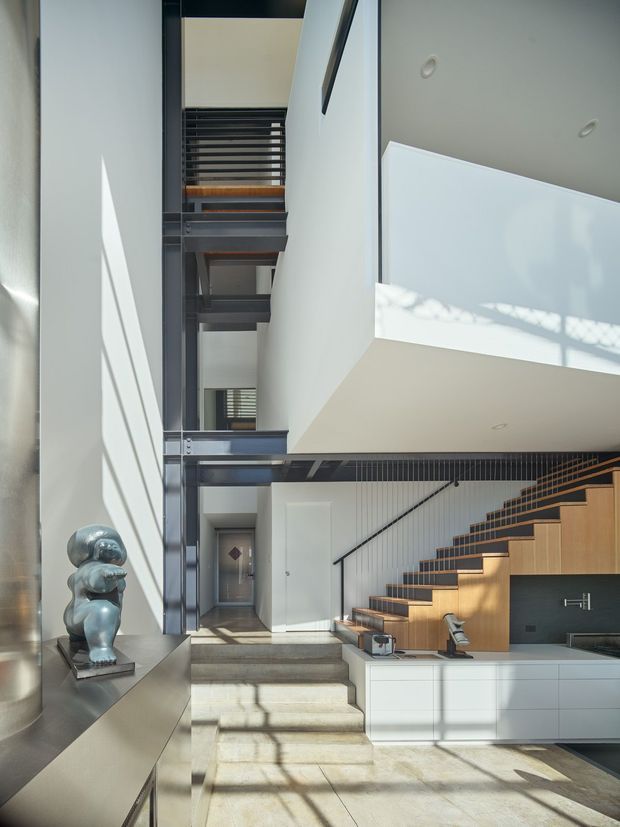 A bright, double-height contemporary interior combining a kitchen, stairway and hallway. The space features a sculptural open-riser timber staircase with dark metal stringers and balustrade, white walls, extensive glazing or reflective surfaces, and white built-in kitchen cabinetry with a dark benchtop. Concrete or stone steps lead through the open-plan area, while strong sunlight creates graphic shadows. A metallic sculptural object sits on a built-in ledge along the left wall.