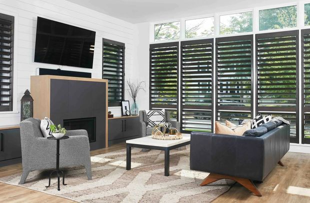 Bright contemporary living room with white horizontal wall panelling, a dark built-in fireplace and television wall, grey armchair and sofa, low square coffee table, patterned rug, and extensive black louvred glazing overlooking greenery. Natural timber flooring and abundant filtered daylight create a refined indoor-outdoor feel.