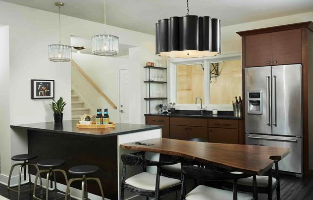A contemporary kitchen with a dark island and breakfast bar, warm wood cabinetry, stainless-steel appliances, open shelving, and a separate timber dining table with black chairs. Pendant lights hang above the island and dining area, while a stairway is visible beyond the kitchen.