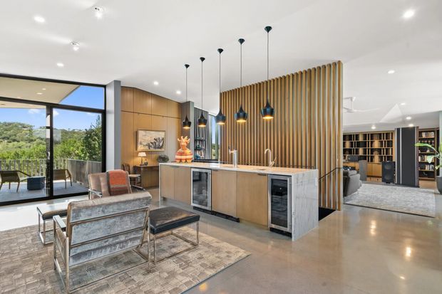 Open-plan contemporary kitchen and living area with a large central island, timber cabinetry, marble-look end panels, a vertical timber batten feature screen, and multiple pendant lights. Floor-to-ceiling glazing opens to a balcony with leafy views. A seating area and extensive built-in shelving are visible beyond the kitchen.
