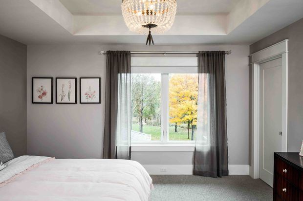A refined bedroom with a partially visible upholstered bed, soft white and blush bedding, dark grey walls, sheer and charcoal curtains, built-in wardrobe cabinetry, framed botanical artwork, and a large central window overlooking trees. A faceted crystal pendant hangs from a recessed tray ceiling, adding an elegant focal point.