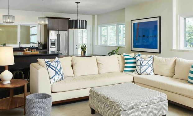Open-plan living room with a large cream sectional sofa and patterned ottoman in the foreground, connected to a compact kitchen with dark cabinetry, stainless-steel appliances, and a peninsula. Large windows provide abundant daylight, while blue and teal accents appear in the cushions and framed artwork. Pendant lights and simple contemporary furnishings complete the space.