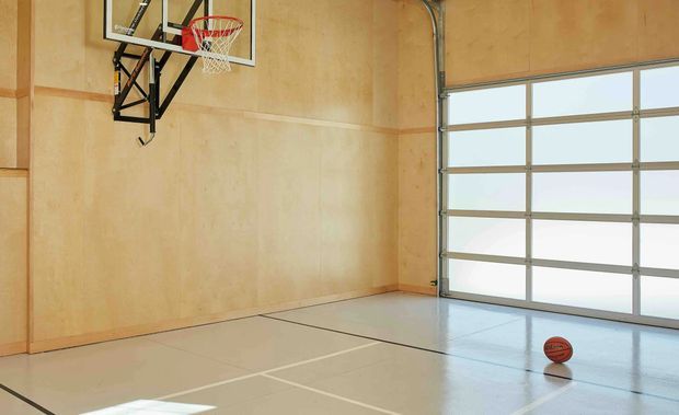 A bright indoor basketball court or home gym with pale timber wall panelling, a mounted basketball hoop, painted court lines on a smooth light floor, and a large translucent sectional garage-style door allowing daylight into the space.