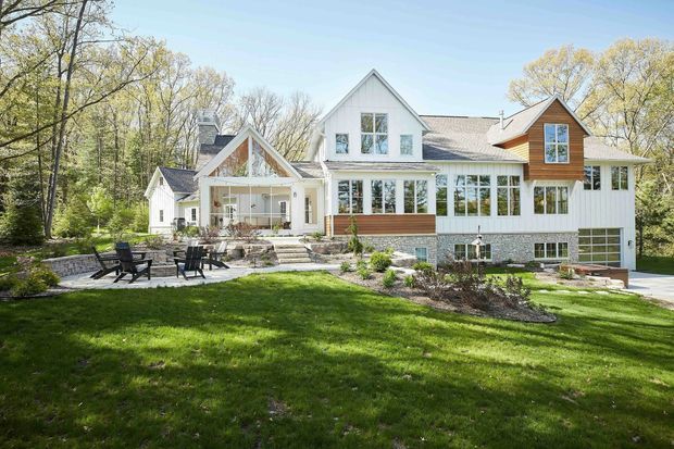 Exterior view of a large multi-gabled suburban house set within a wooded landscaped property. The facade combines white vertical siding, timber-toned horizontal cladding, prominent pitched gables, and extensive white-framed glazing. A raised stone terrace with outdoor seating, steps, retaining walls, and lawn occupies the foreground.