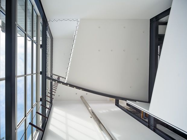 Upward-looking architectural interior view of a bright multi-level stairwell or atrium. Tall black-framed glazing runs along the left side, with open stair landings, dark metal balustrades, and a large white wall and ceiling plane forming a strong geometric composition. The image emphasizes verticality, daylight, and exposed circulation space.