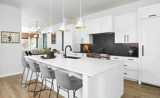 Bright contemporary open-plan kitchen dominated by a large white island with integrated seating for four. White full-height cabinetry, a dark herringbone tile splashback, stainless cooking appliances, timber-look flooring, and three white pendant lights with brass interiors create a clean, refined space. A dining area and windows are visible beyond the island.